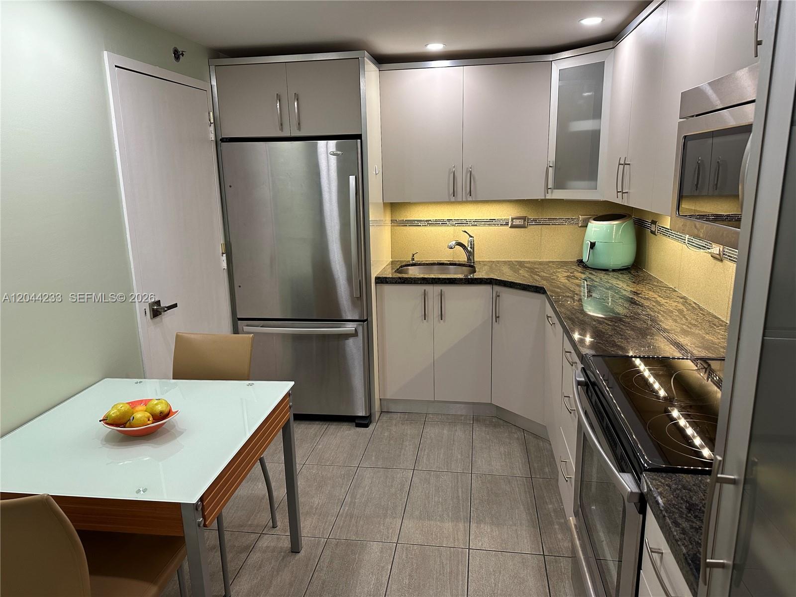 A kitchen features stainless steel appliances, light-colored cabinetry, dark granite countertops, and tile flooring — 6039 Collins Ave # 1205, Miami Beach FL 33140, MIAMI BEACH, FL