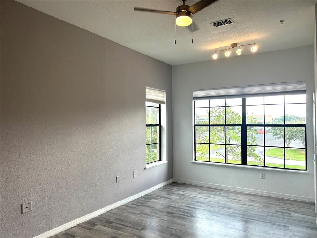 An empty room features gray walls, wood-look flooring, a ceiling fan, and large windows — 6137 METROWEST BOULEVARD UNIT 307, ORLANDO, FL