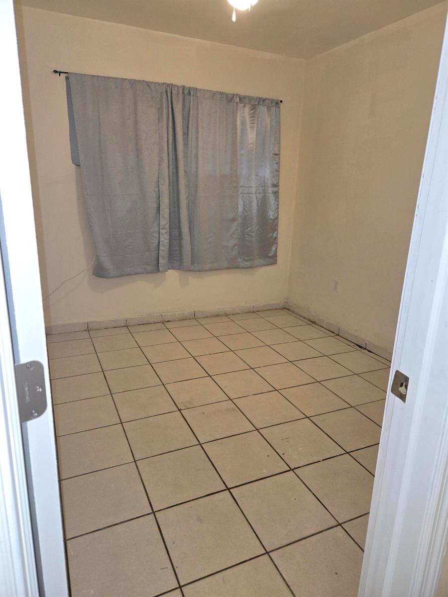 An empty bedroom features light-colored tile flooring, beige walls, and light blue curtains — 1560 NE 111th Street # 3, Miami FL 33161, MIAMI, FL