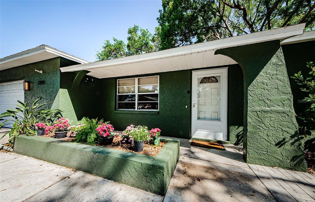 A green house exterior features a front garden bed with pink flowers and a white door — 7241 56TH AVENUE N, ST PETERSBURG, FL