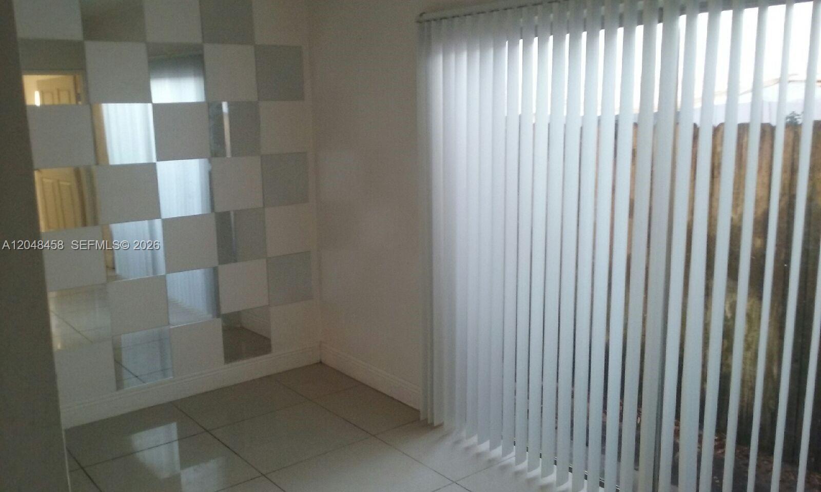 A room features light-colored tile flooring, a mirrored accent wall, and vertical window blinds — NORTH MIAMI BEACH, FL
