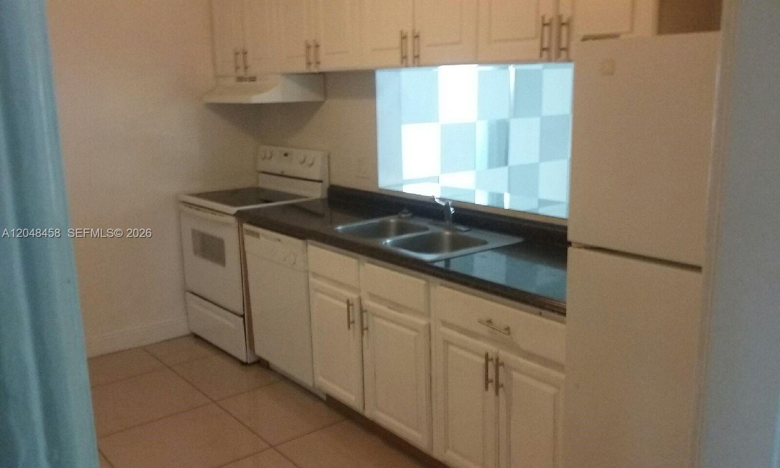 A kitchen features white cabinets, a dark countertop, a double sink, and white appliances — NORTH MIAMI BEACH, FL