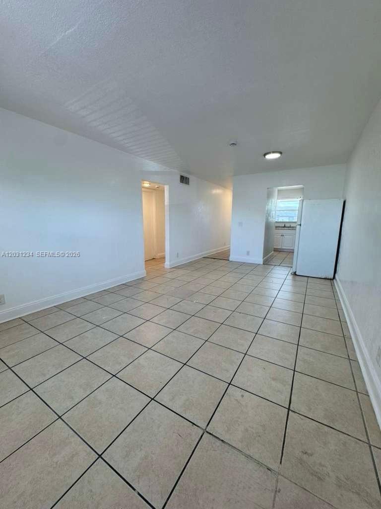An empty room with light-colored tile flooring leads to a kitchen and a hallway — RIVIERA BEACH, FL