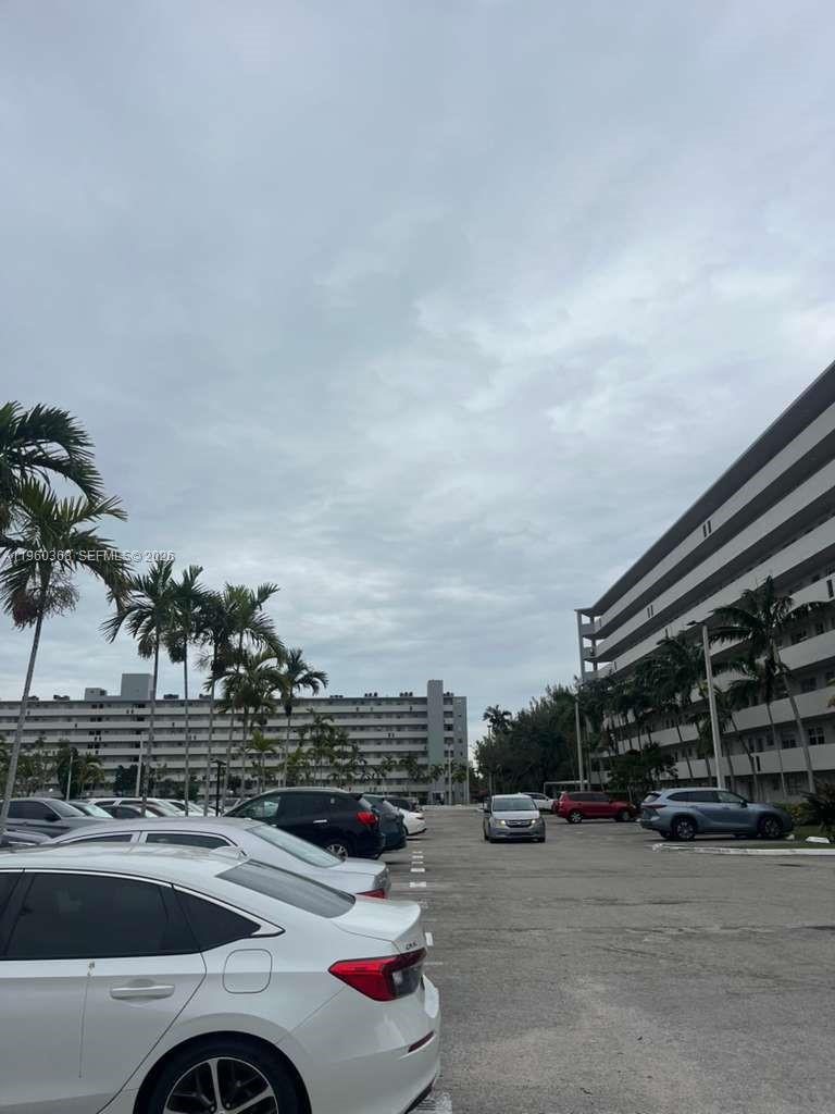 An outdoor parking lot is situated between two large residential apartment buildings under a cloudy sky — 1750 NE 191st St # 1, Miami FL 33179, MIAMI, FL