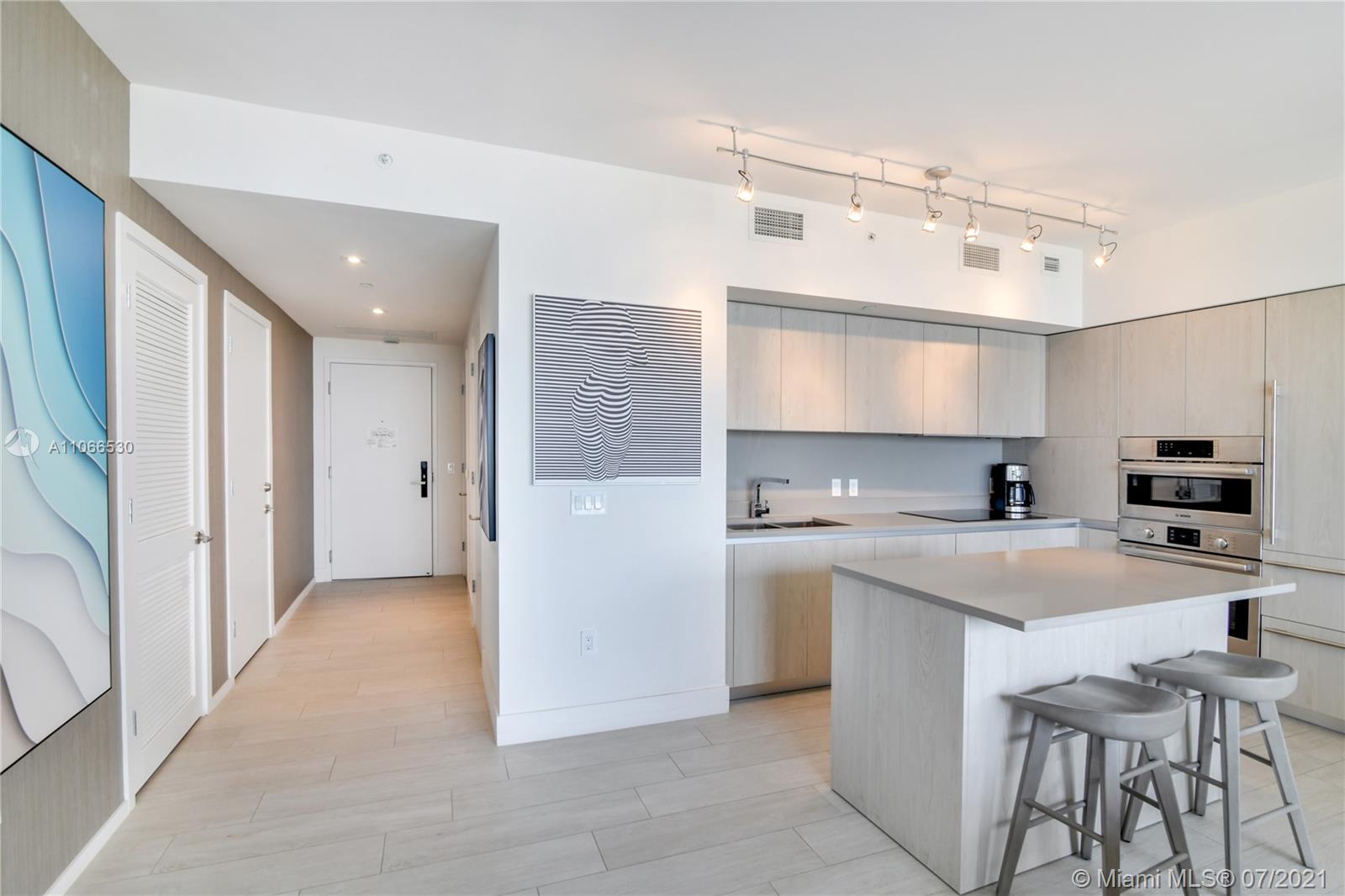 A modern kitchen features light wood cabinetry, a central island with stools, and white walls — 4010 S Ocean Dr # R1406, Hollywood FL 33019, HOLLYWOOD, FL