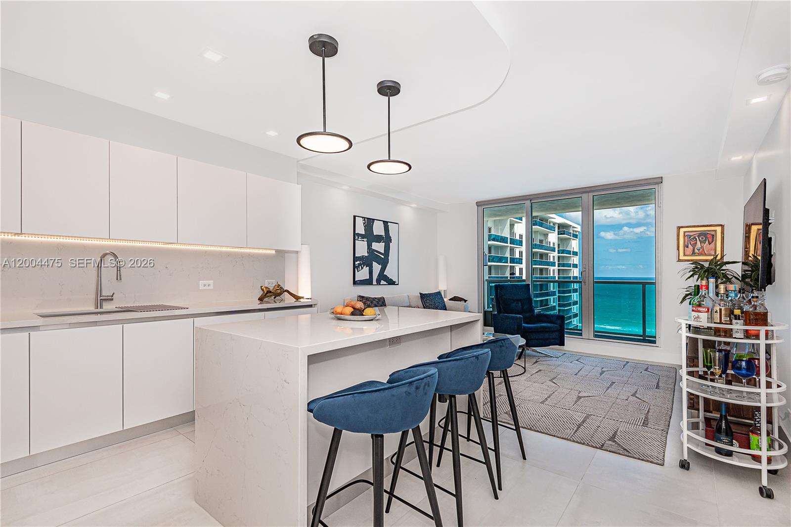 A modern kitchen and living area features a white island, blue stools, and ocean views — 2301 Collins Ave # 1440, Miami Beach FL 33139, MIAMI BEACH, FL