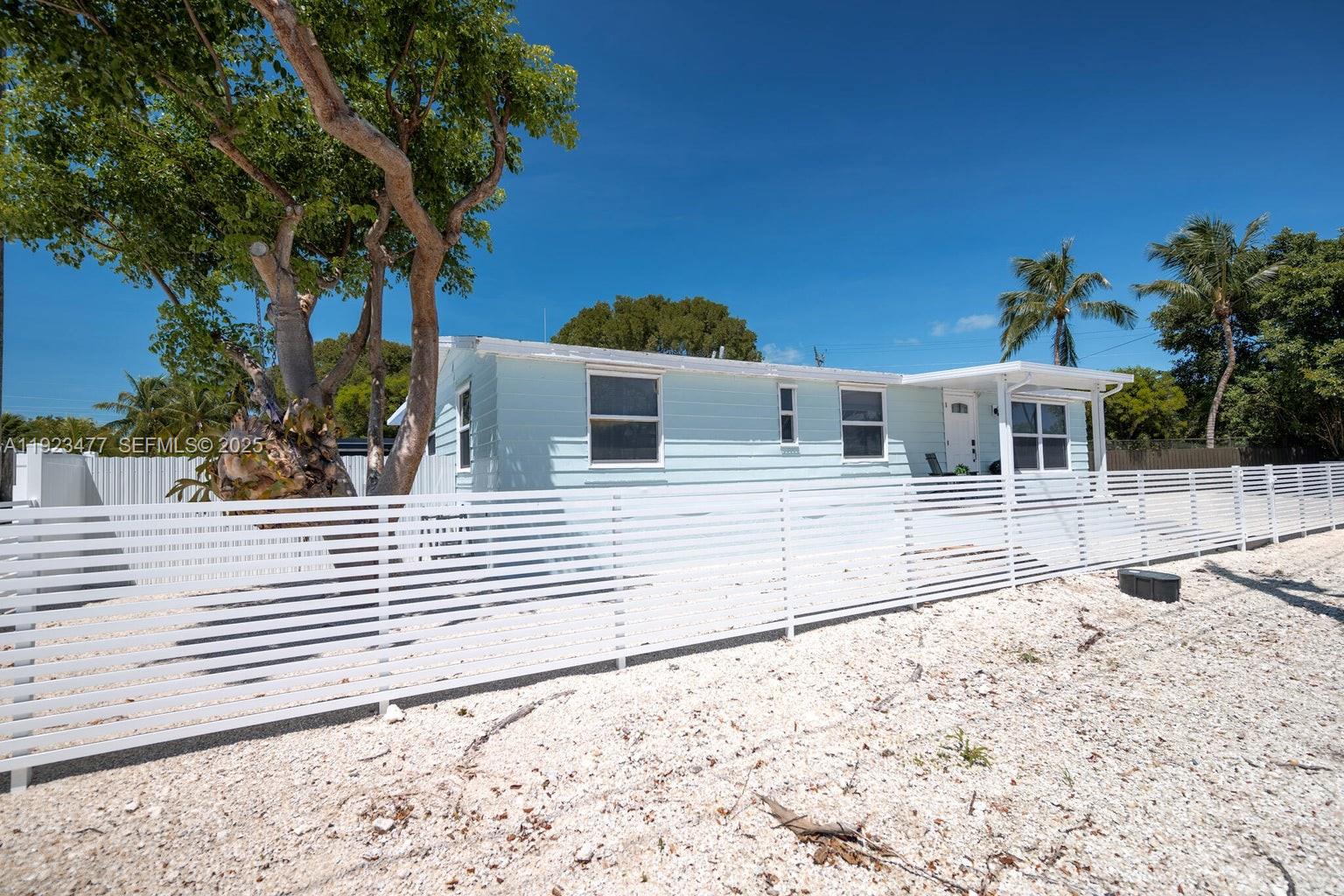 A light blue single-story house sits behind a white horizontal slat fence on a gravel lot — 212 Buttonwood Ln, Key Largo FL 33070, KEY LARGO, FL