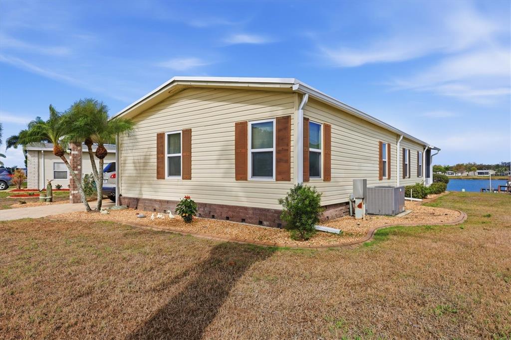 A light-yellow mobile home with brown shutters sits on a gravel lot near water — 6085 SHEARWATER DRIVE, ENGLEWOOD, FL