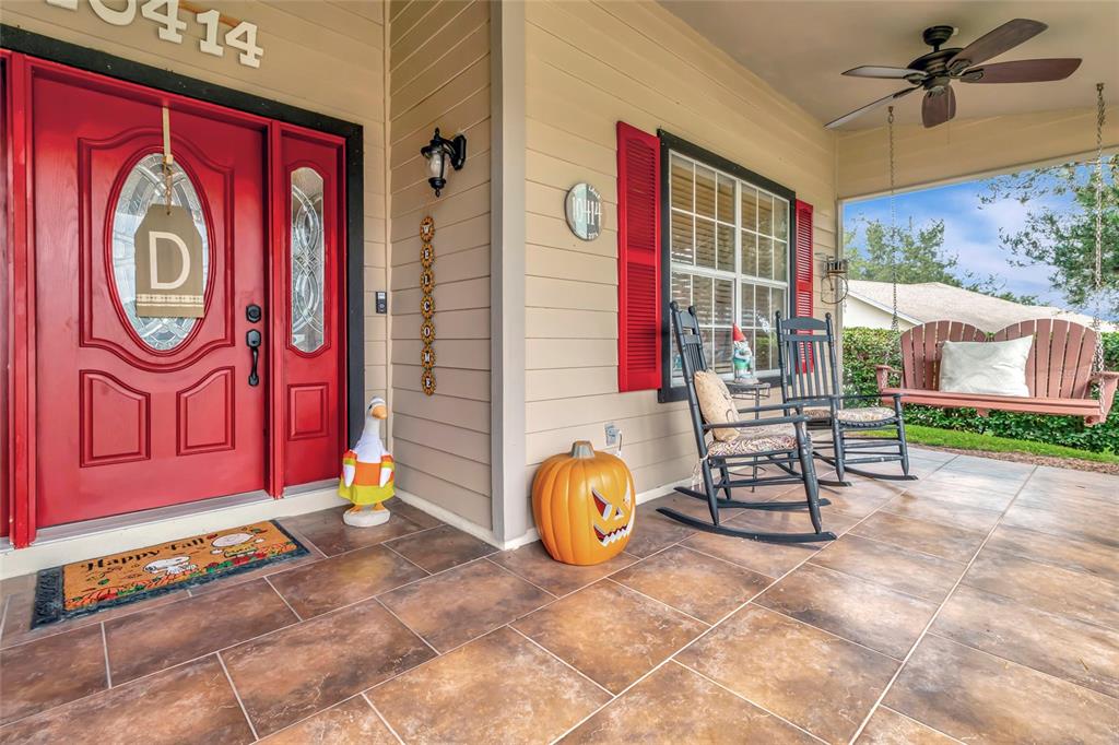 A covered front porch features tiled flooring, a red door, rocking chairs, and seasonal decor — 10414 MESA LANE, CLERMONT, FL