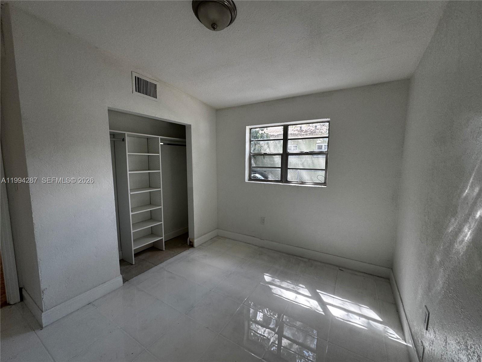 A small, empty bedroom features white walls, tiled floors, a window, and a built-in closet — 1950 Washington Ave # 2, Opa-Locka FL 33054, OPA-LOCKA, FL