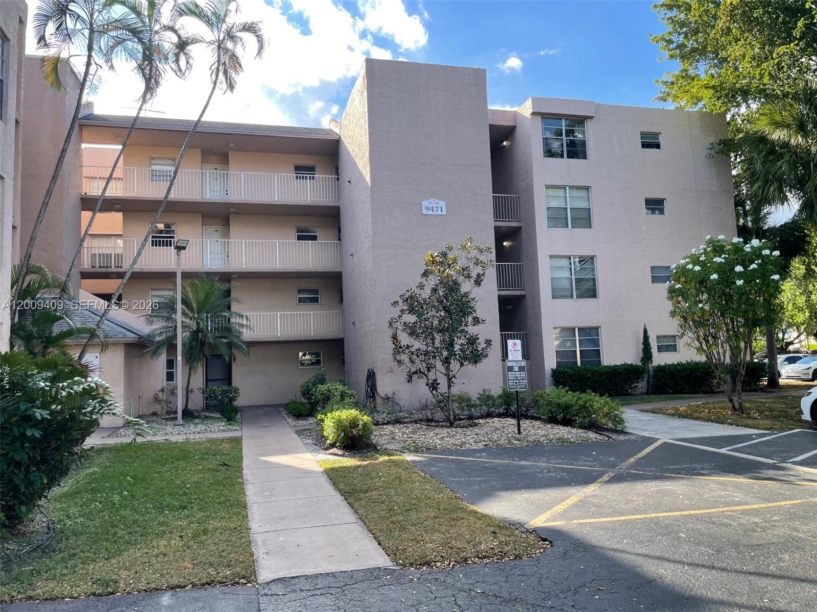 A multi-story apartment building features beige stucco walls, open balconies, and a paved parking area — 9471 Evergreen Pl # 401, Davie FL 33324, DAVIE, FL