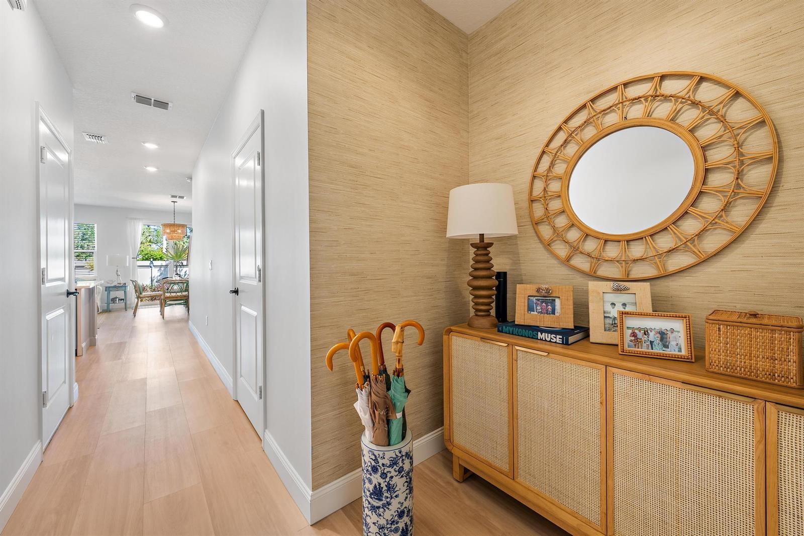 A hallway features light wood flooring, textured wallpaper, a rattan console table, and a round mirror — 327 SE Halifax Lane, Stuart FL 34994, STUART, FL