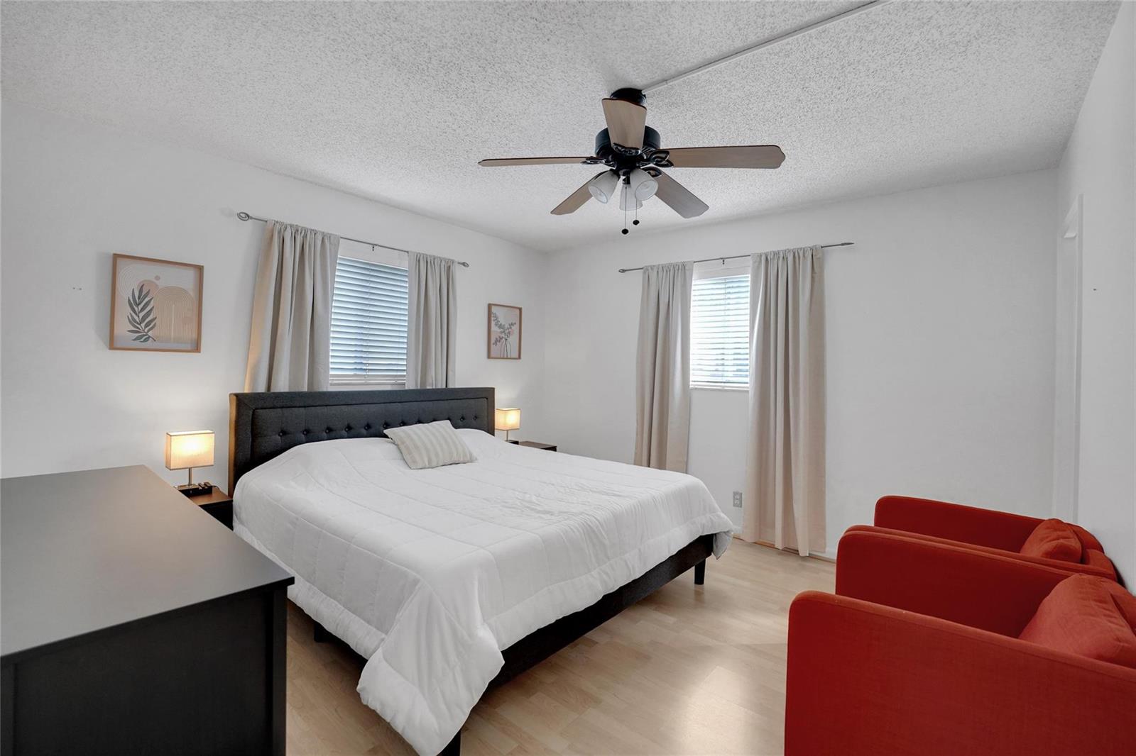A bedroom features a bed with a dark headboard, two red chairs, and light flooring — 257 S Cypress Road # 401, Pompano Beach FL 33060, POMPANO BEACH, FL