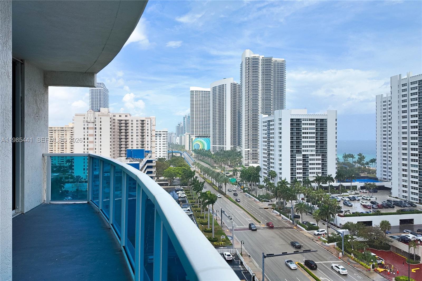 A balcony view overlooks a multi-lane road lined with palm trees and tall residential buildings — 1945 S Ocean Dr # 1114, Hallandale Beach FL 33009, HALLANDALE BEACH, FL