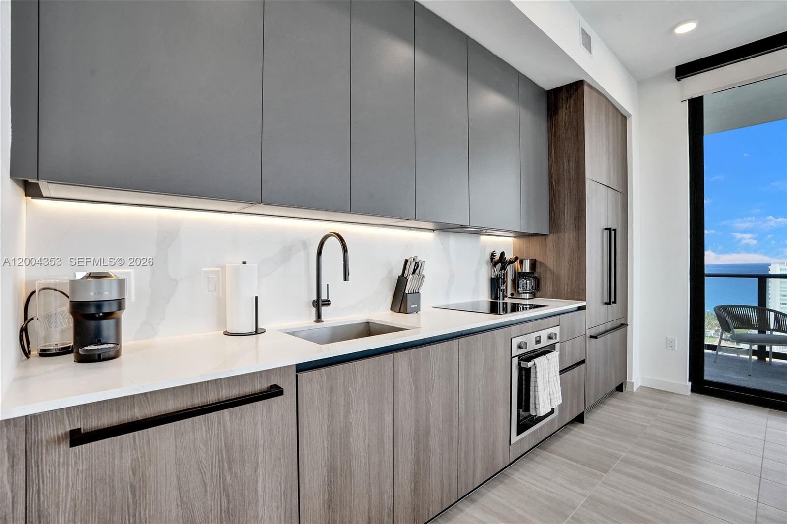 A modern kitchen features gray upper cabinets, wood-grain lower cabinets, white countertops, and a balcony view — 580 72nd St # 1702, Miami Beach FL 33141, MIAMI BEACH, FL