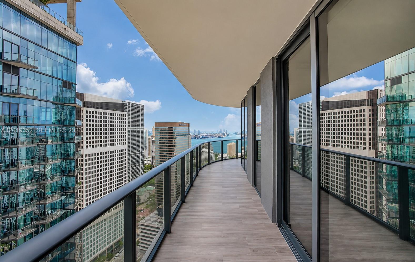 A curved balcony with wood-look tile flooring overlooks a city skyline and a body of water — 801 S Miami Ave # 3905, Miami FL 33130, MIAMI, FL