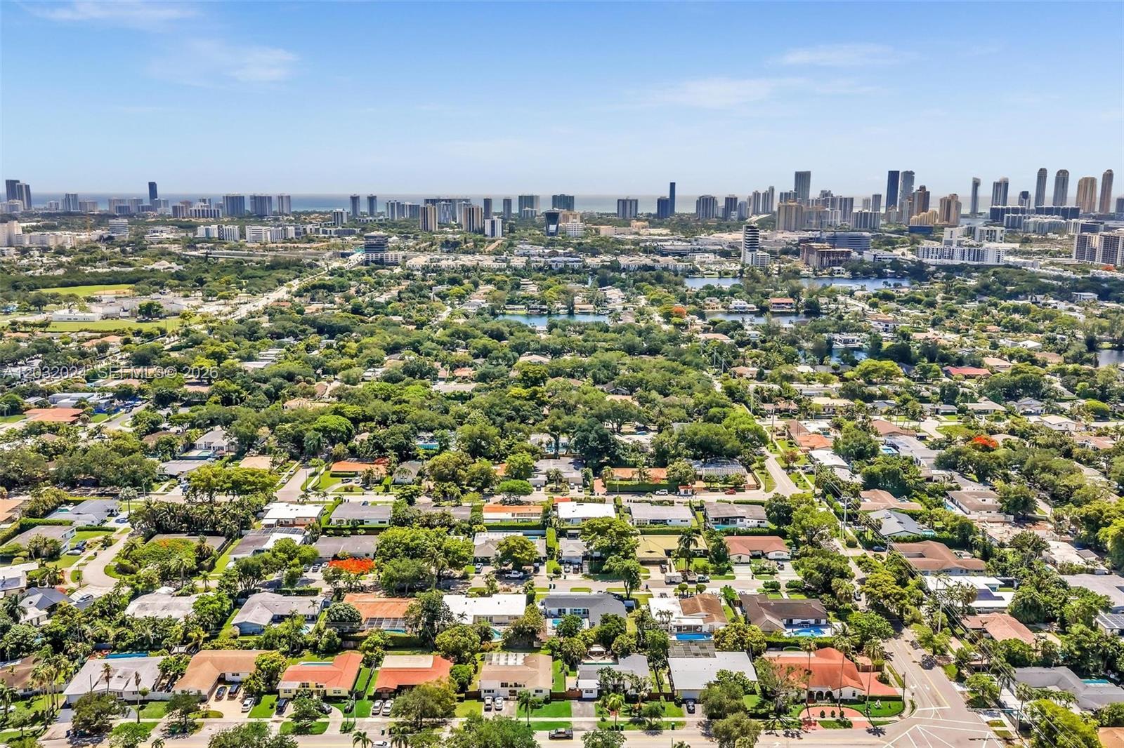 An aerial view shows a residential neighborhood with lush trees and a distant city skyline — 19901 Highland Lakes Blvd, Miami FL 33179, MIAMI, FL