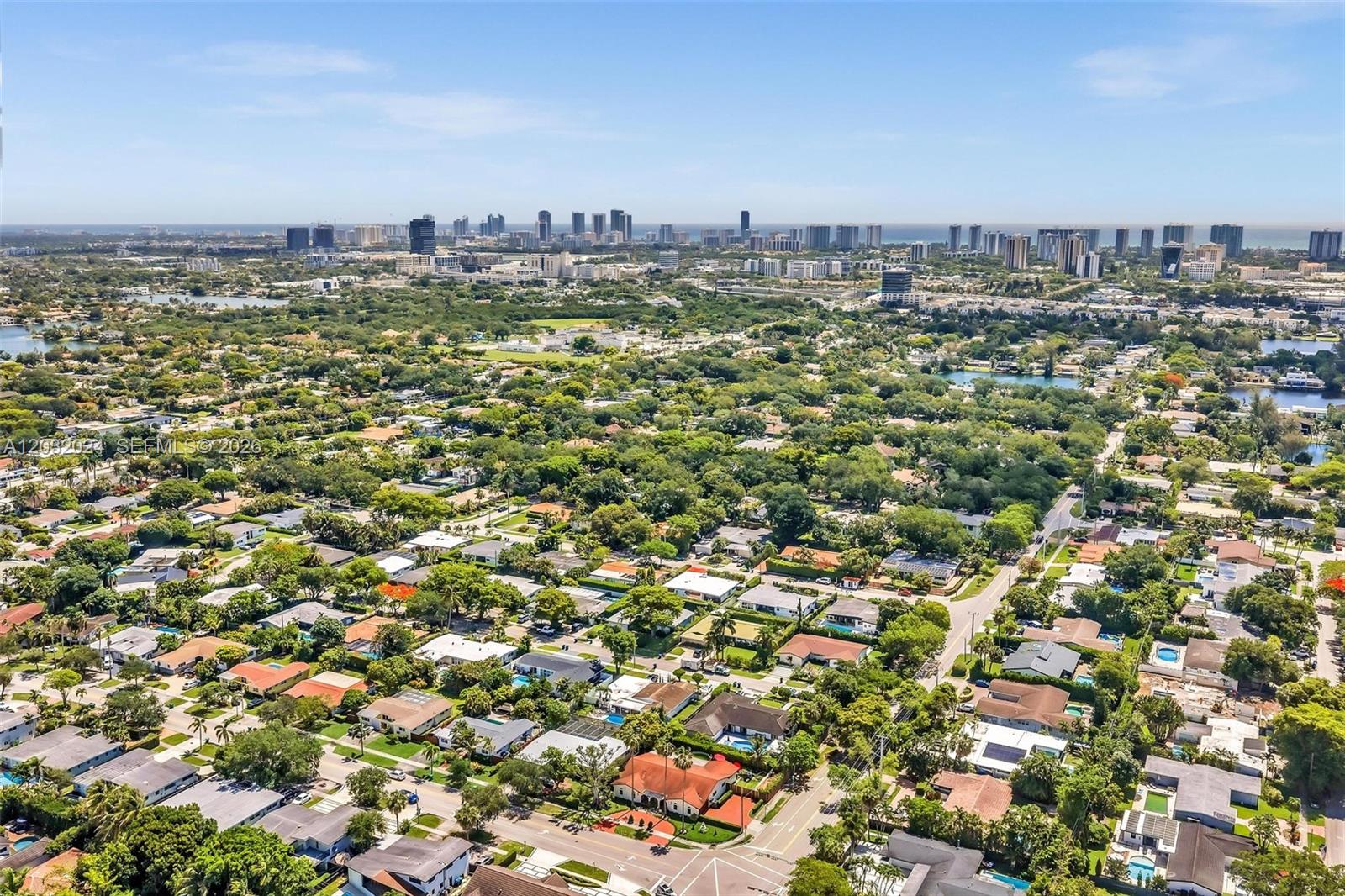 An aerial view shows a suburban neighborhood with many trees and a distant city skyline — 19901 Highland Lakes Blvd, Miami FL 33179, MIAMI, FL
