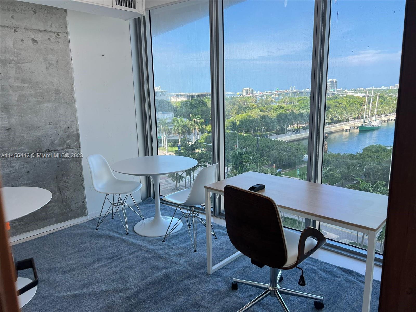 A room with a desk, a small table, and floor-to-ceiling windows overlooking a waterfront — 888 Biscayne  Blvd, Miami FL 33132, MIAMI, FL