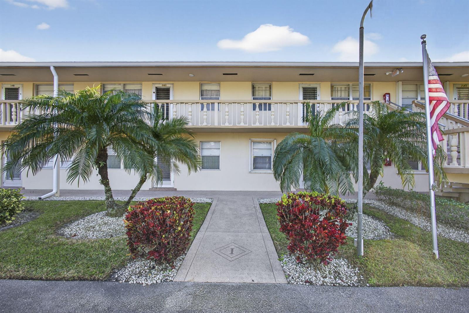 A two-story apartment building exterior features a concrete walkway, palm trees, and an American flag — 149 Hastings I, West Palm Beach FL 33417, WEST PALM BEACH, FL