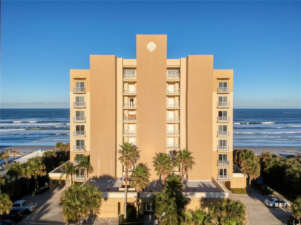 An exterior view of a multi-story tan beachfront condominium building with palm trees and parking — 1705 S ATLANTIC AVENUE UNIT 804, NEW SMYRNA BEACH, FL