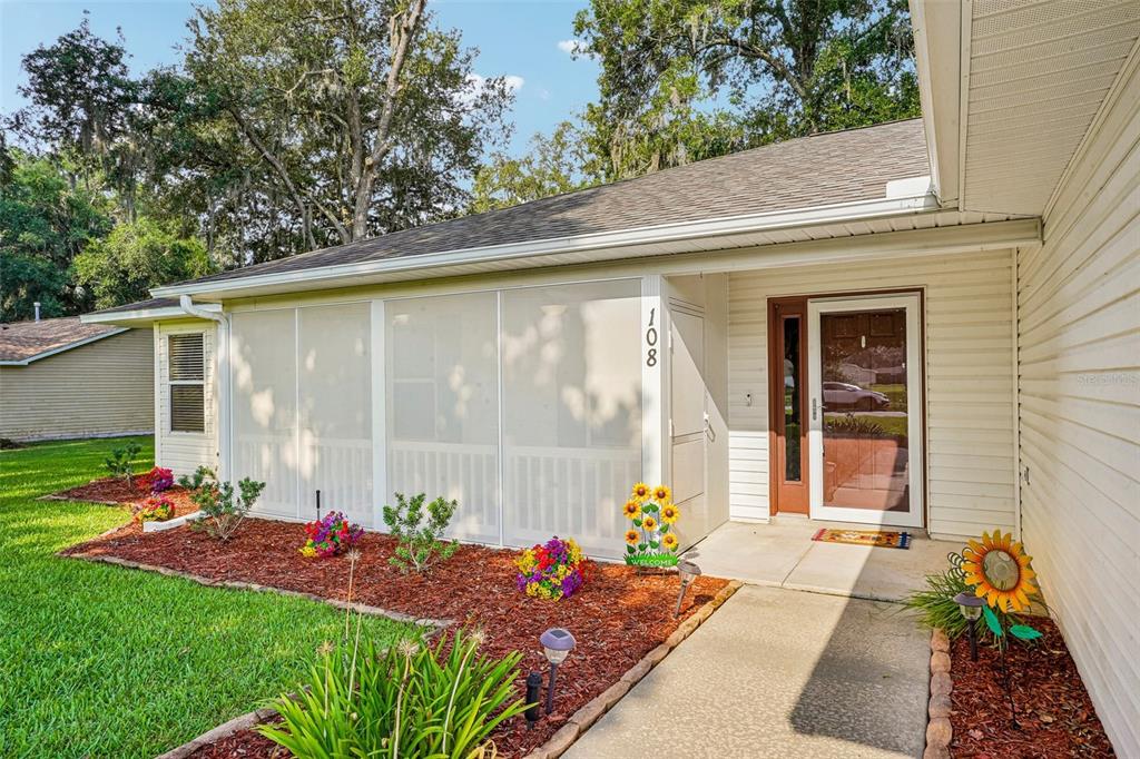 A single-story house exterior featuring a screened-in porch, a concrete walkway, and landscaped flower beds — 108 CHERRY BLOSSOM LANE, LADY LAKE, FL