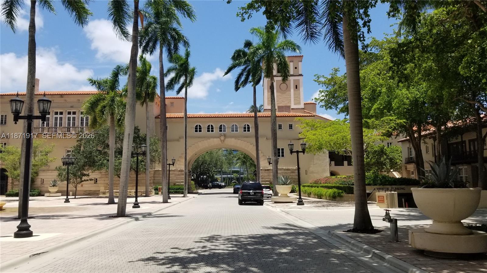 A paved driveway leads toward a large stone archway connecting two Mediterranean-style buildings — 888 S Douglas Rd # 1213, Coral Gables FL 33134, CORAL GABLES, FL