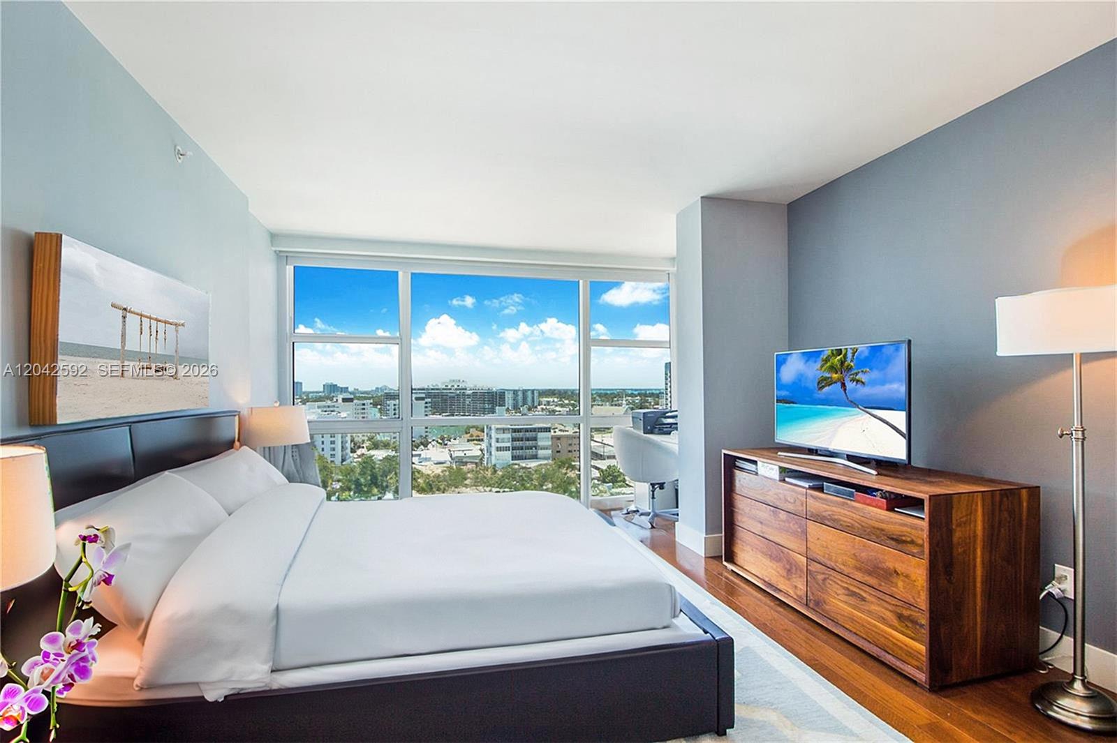 A bedroom features a bed, wooden dresser with a television, and floor-to-ceiling windows with city views — 6801 Collins Ave # 1101, Miami Beach FL 33141, MIAMI BEACH, FL