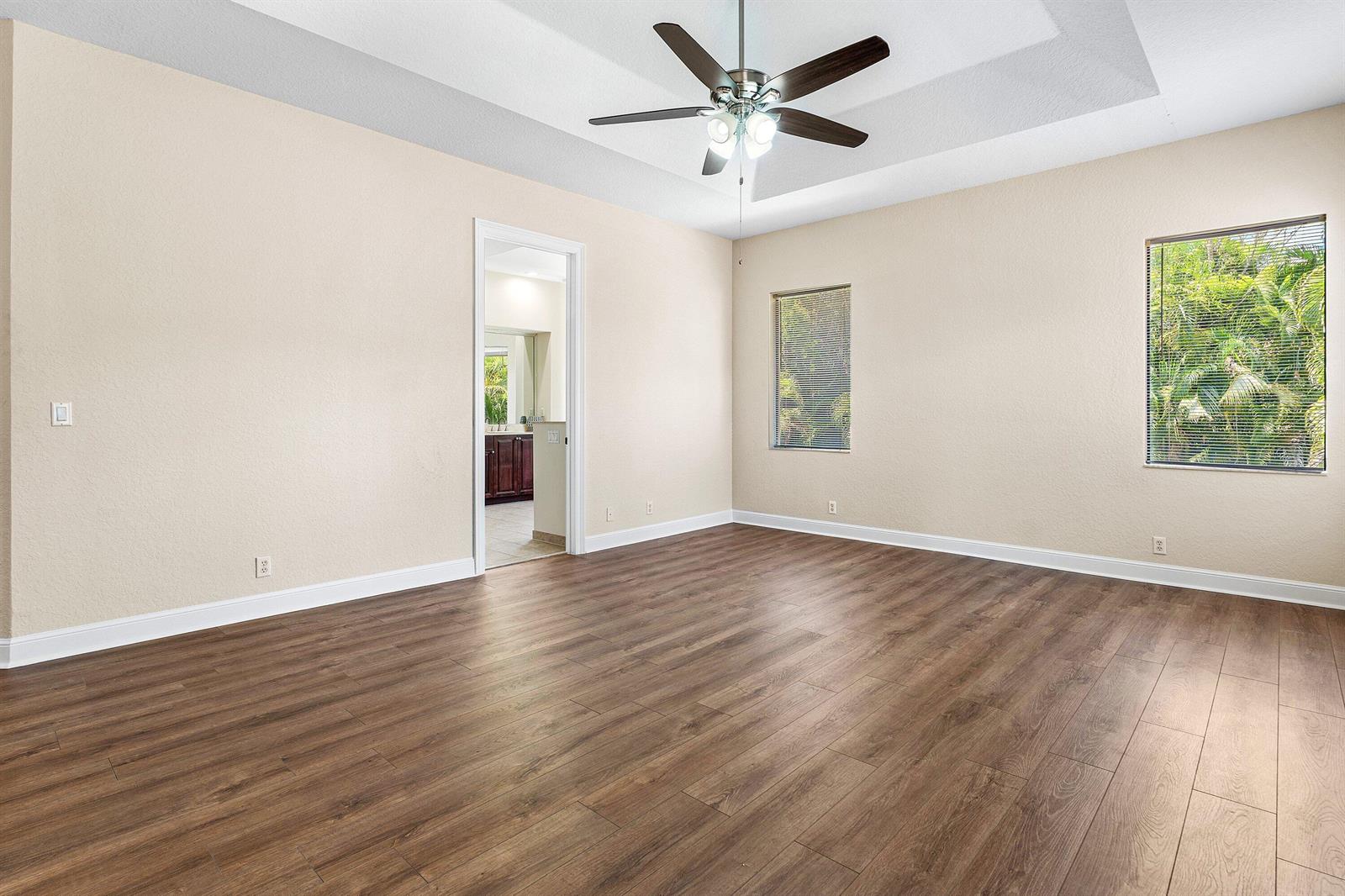 An empty bedroom with wood-look flooring, a ceiling fan, and two windows overlooking greenery — 10293 Medicis Place, Wellington FL 33449, WELLINGTON, FL