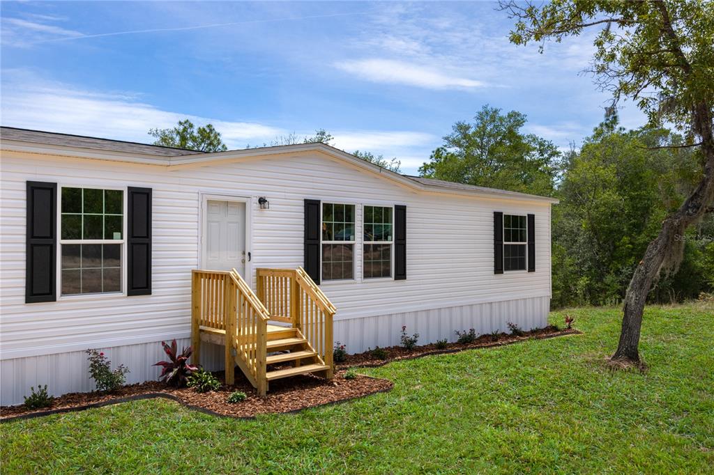 A white manufactured home exterior features black shutters, a wooden staircase, and a landscaped yard — 119 LONDON AVENUE, INTERLACHEN, FL