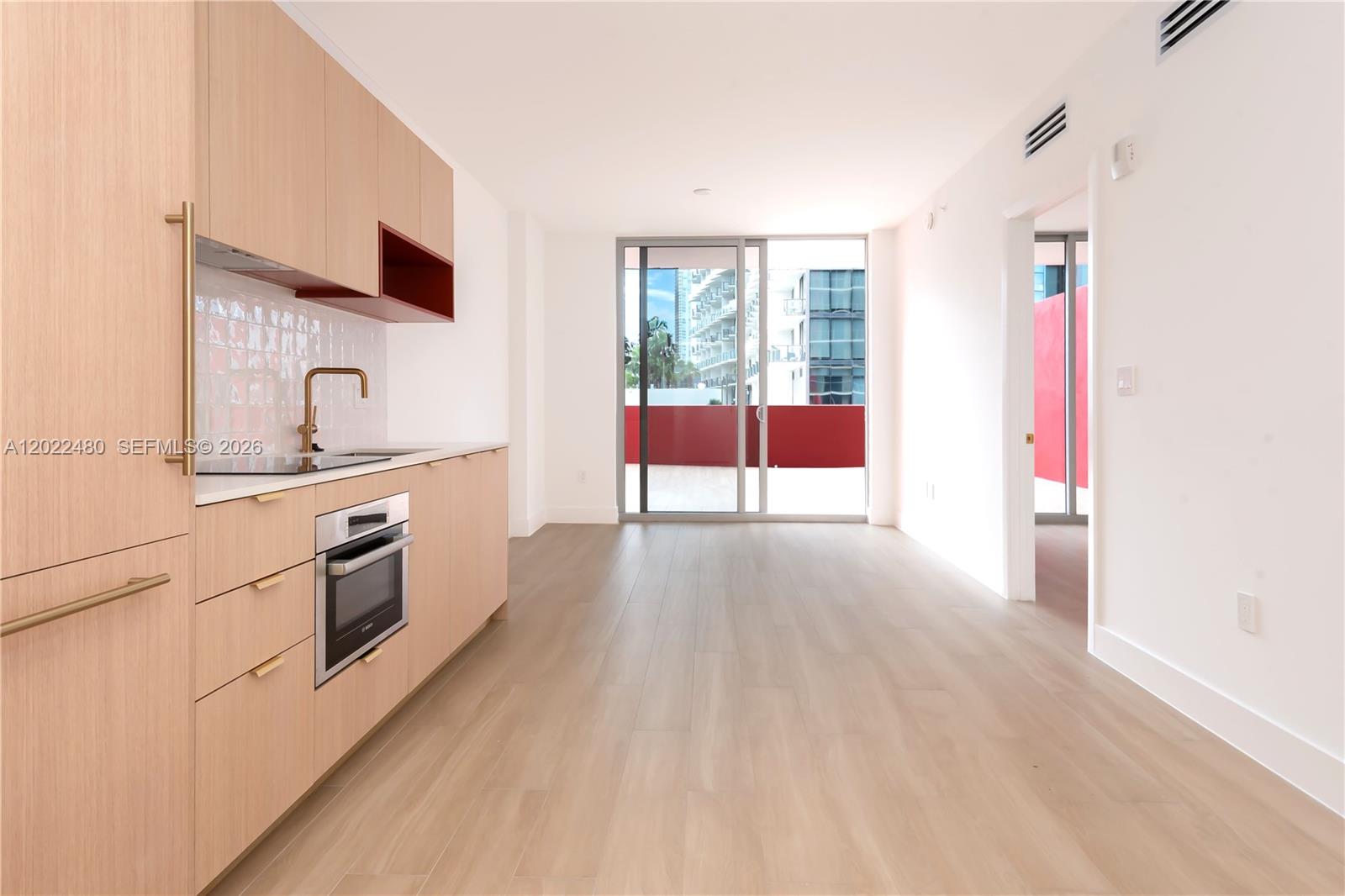 A modern kitchen with light wood cabinets and a red accent shelf faces a balcony — 90 NE 32 # 608, Miami FL 33137, MIAMI, FL