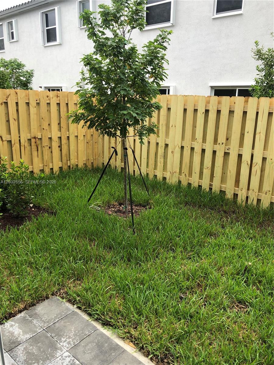 A young tree stands in a grassy backyard enclosed by a light-colored wooden fence — 2610 SE 13th St, Homestead FL 33035, HOMESTEAD, FL