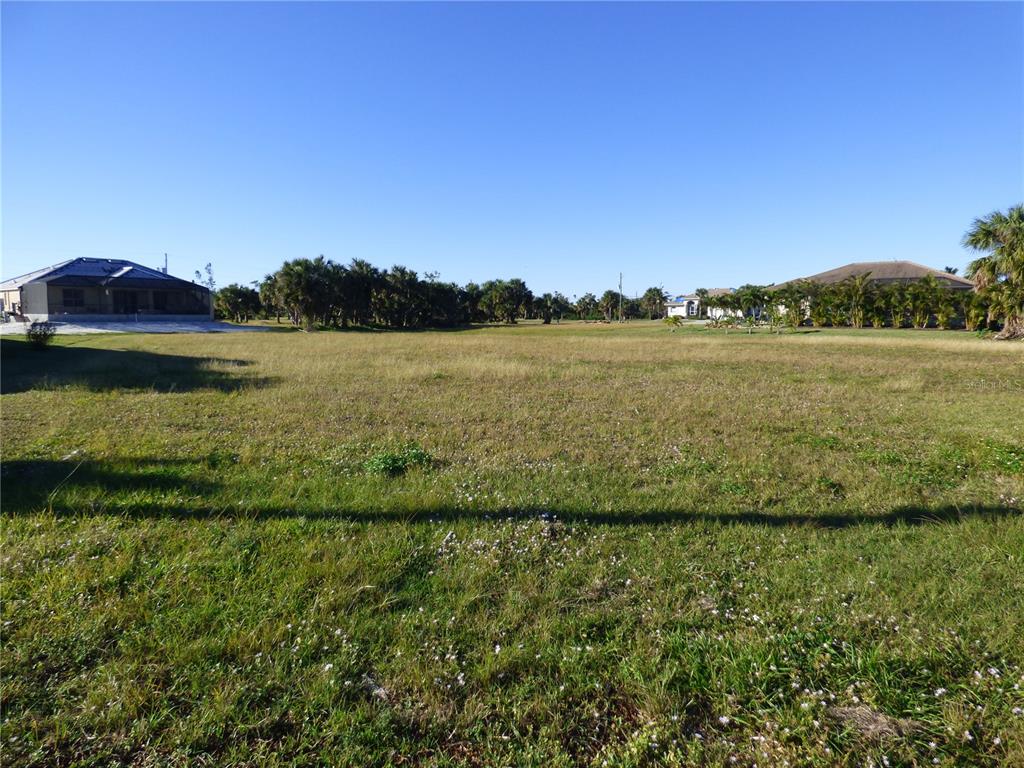 A large, flat, grassy vacant lot sits between two residential houses under a clear sky — 24370 GIJON LANE, PUNTA GORDA, FL