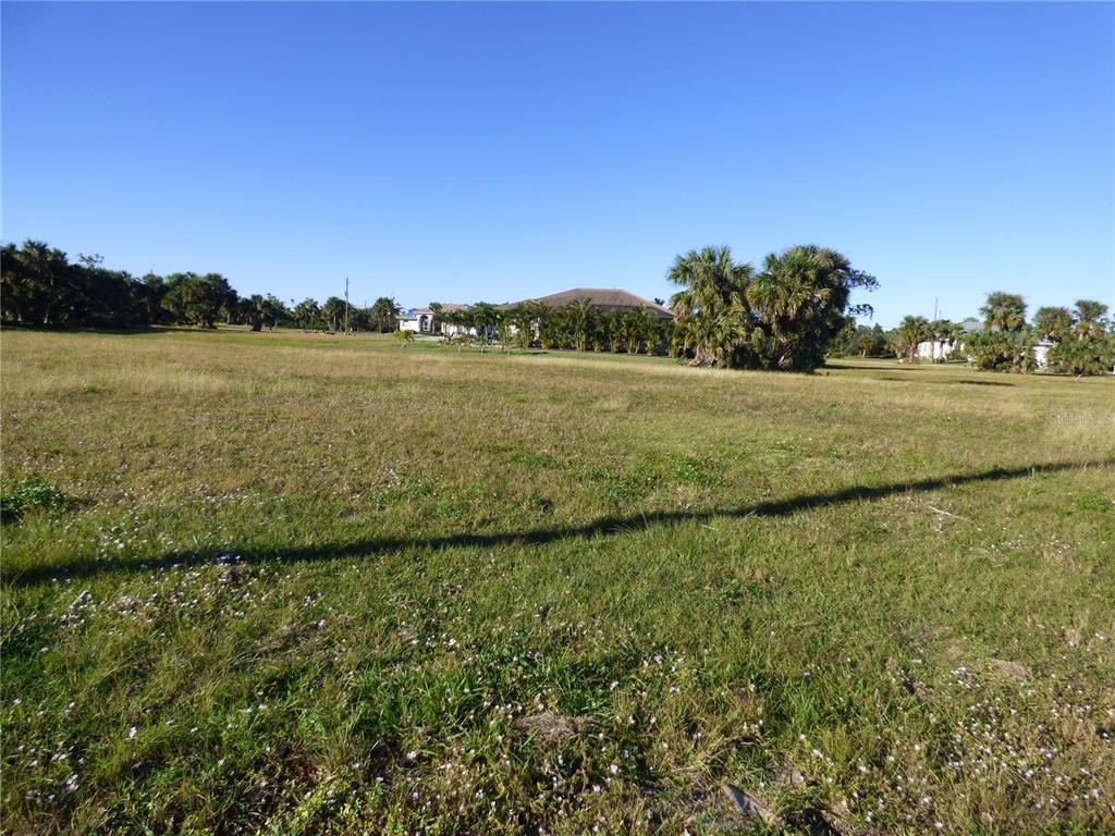 A wide, grassy vacant lot under a clear blue sky with distant trees and houses — 24370 GIJON LANE, PUNTA GORDA, FL