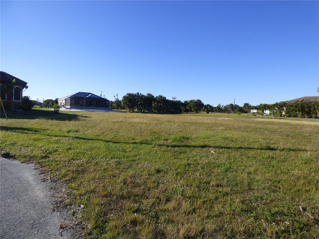 A vacant, grassy residential lot is shown with neighboring houses and trees in the background — 24370 GIJON LANE, PUNTA GORDA, FL