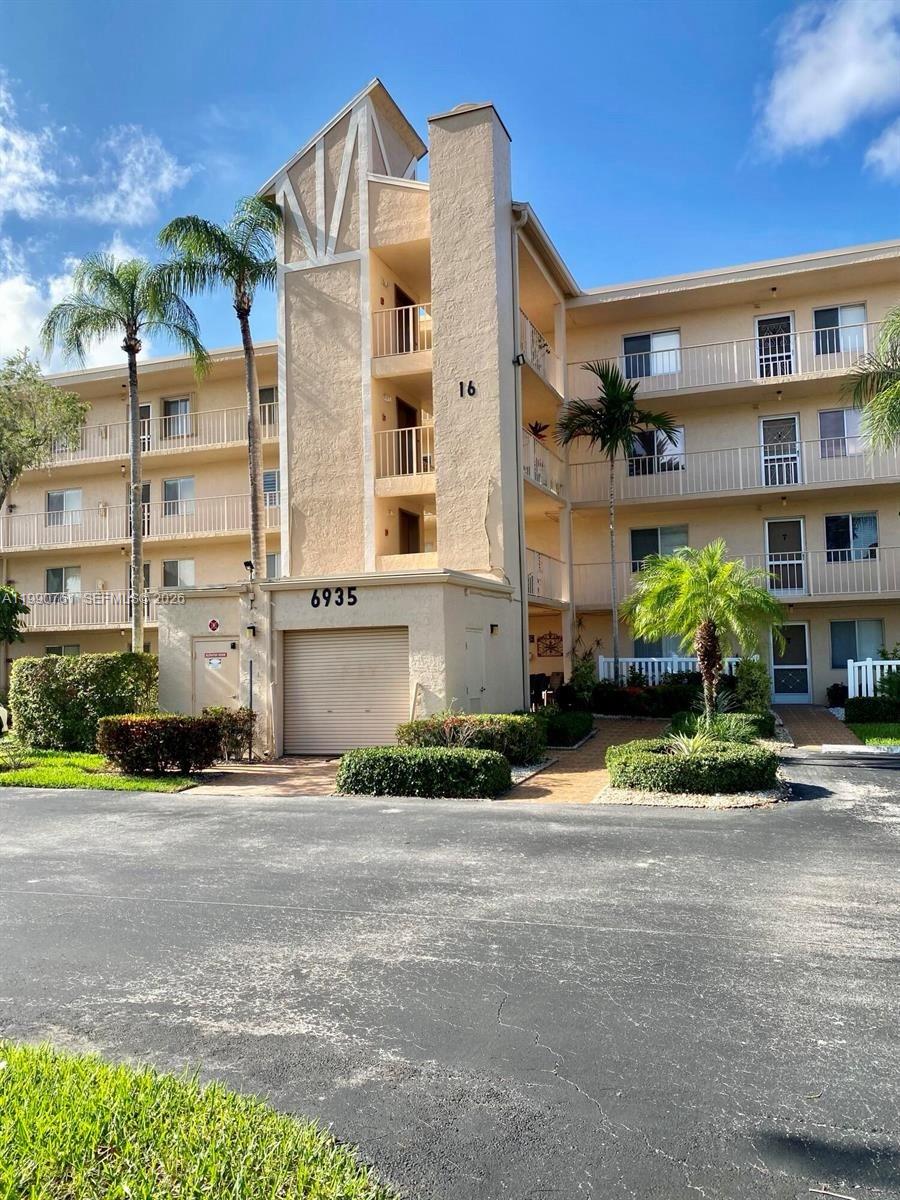 An exterior view of a multi-story apartment building with a garage and palm trees — 6935 Huntington Ln # 108, Delray Beach FL 33446, DELRAY BEACH, FL