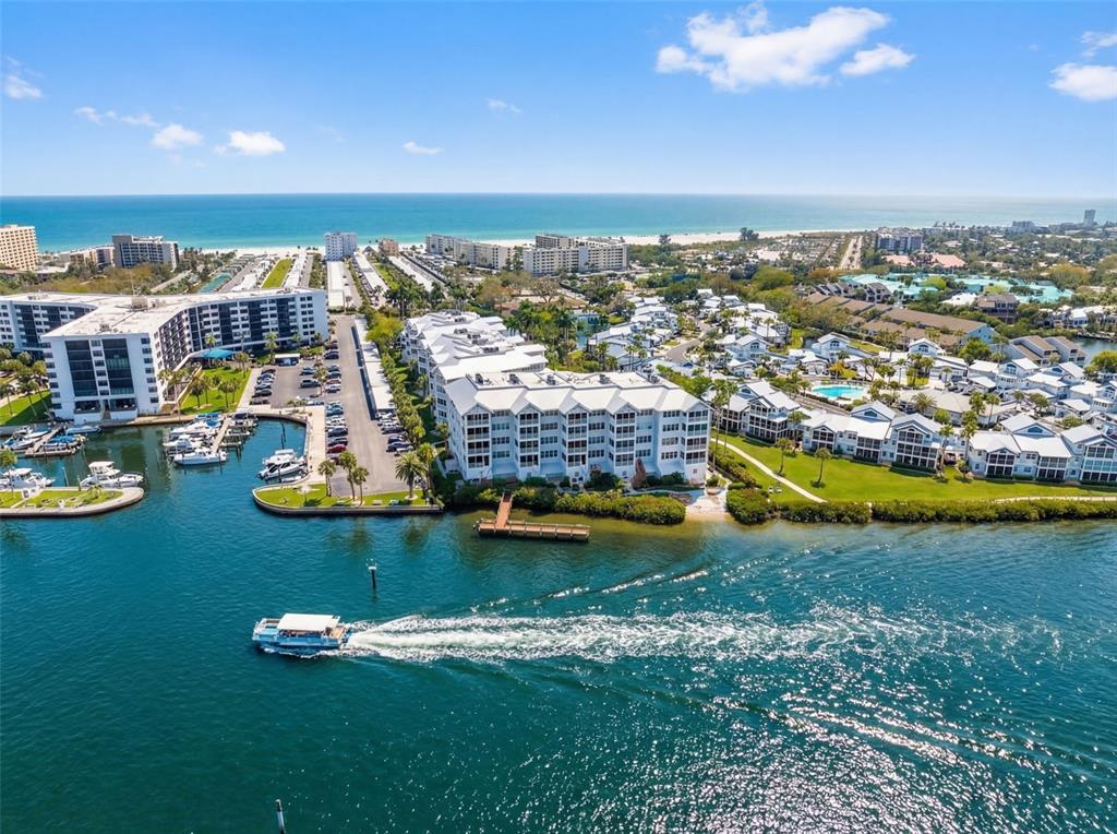 An aerial view shows waterfront condominium buildings, a boat in the canal, and the ocean — 1280 DOLPHIN BAY WAY UNIT 402, SARASOTA, FL