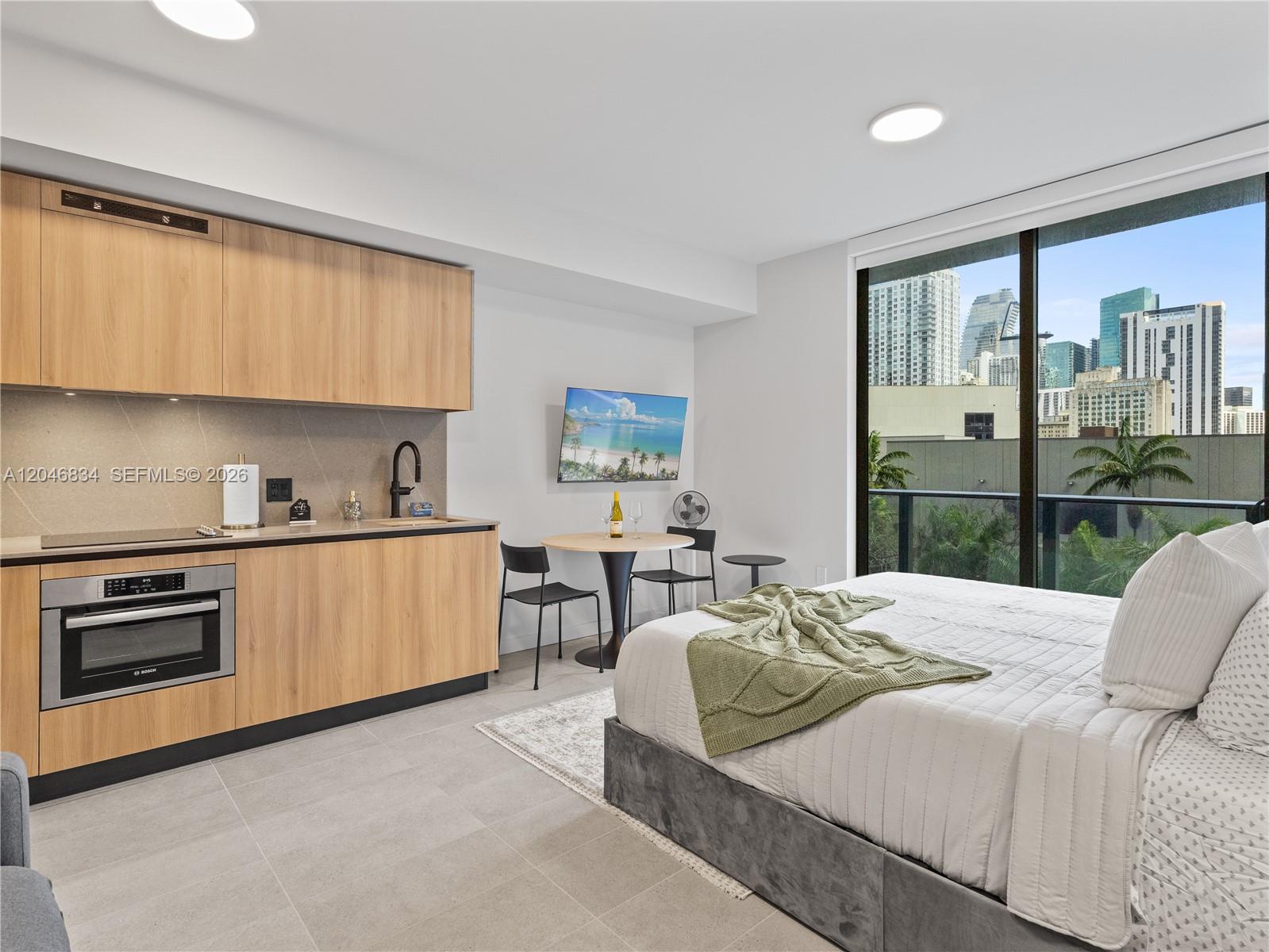 A studio apartment features a kitchenette, a bed, and a balcony overlooking city buildings — 121 NE 5th St # 504, Miami FL 33132, MIAMI, FL