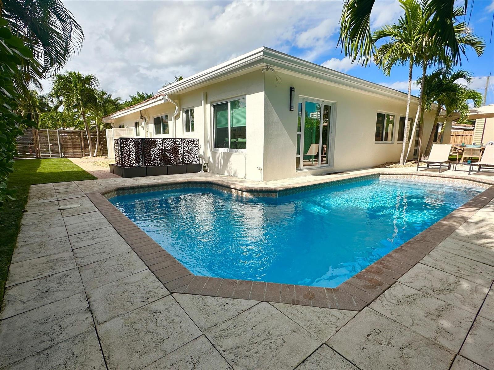 A backyard features a swimming pool with a brick border next to a white house — 1820 Bel Air Avenue, Pompano Beach FL 33062, POMPANO BEACH, FL