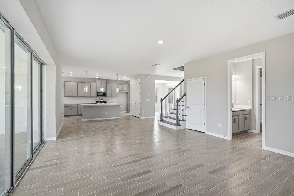 An open-concept living area features light wood-look tile flooring, a kitchen with gray cabinets, and stairs — 1678 TRAILHEAD LANE, ZEPHYRHILLS, FL