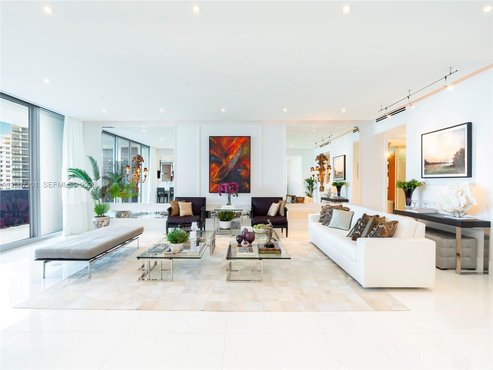 A modern living room features a white sofa, glass coffee tables, and large wall mirrors — 5959 Collins Ave # 906, Miami Beach FL 33140, MIAMI BEACH, FL