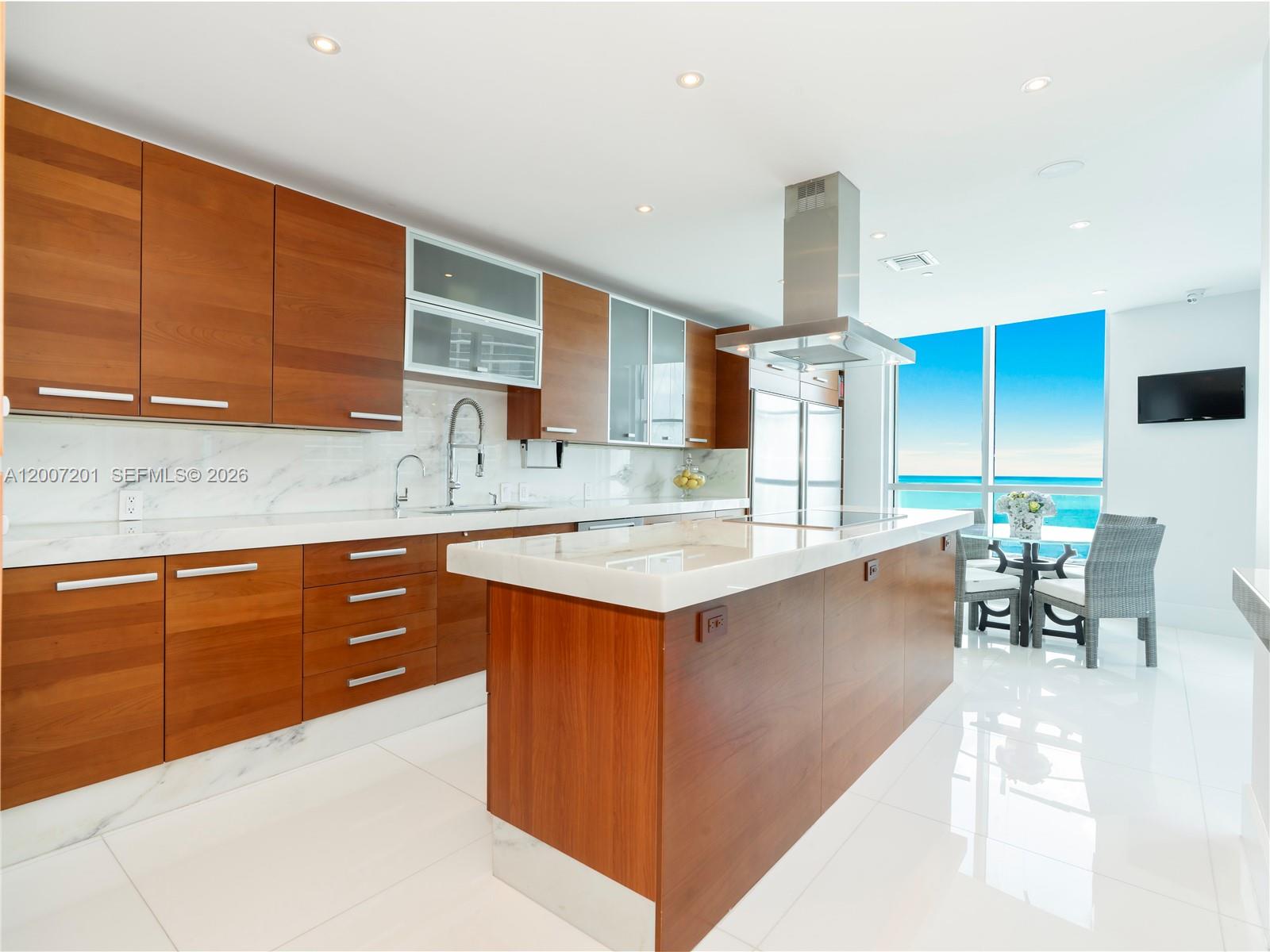A modern kitchen features wood cabinetry, white marble countertops, and an island overlooking the ocean — 5959 Collins Ave # 906, Miami Beach FL 33140, MIAMI BEACH, FL