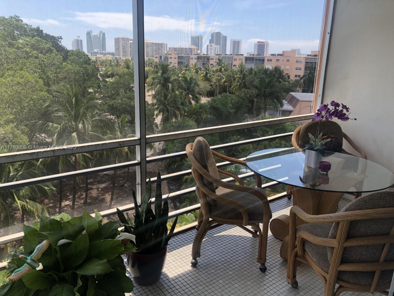 A balcony with a glass-topped table, wicker chairs, and a view of trees and buildings — 1000 NE 14th Ave # 605, Hallandale Beach FL 33009, HALLANDALE BEACH, FL