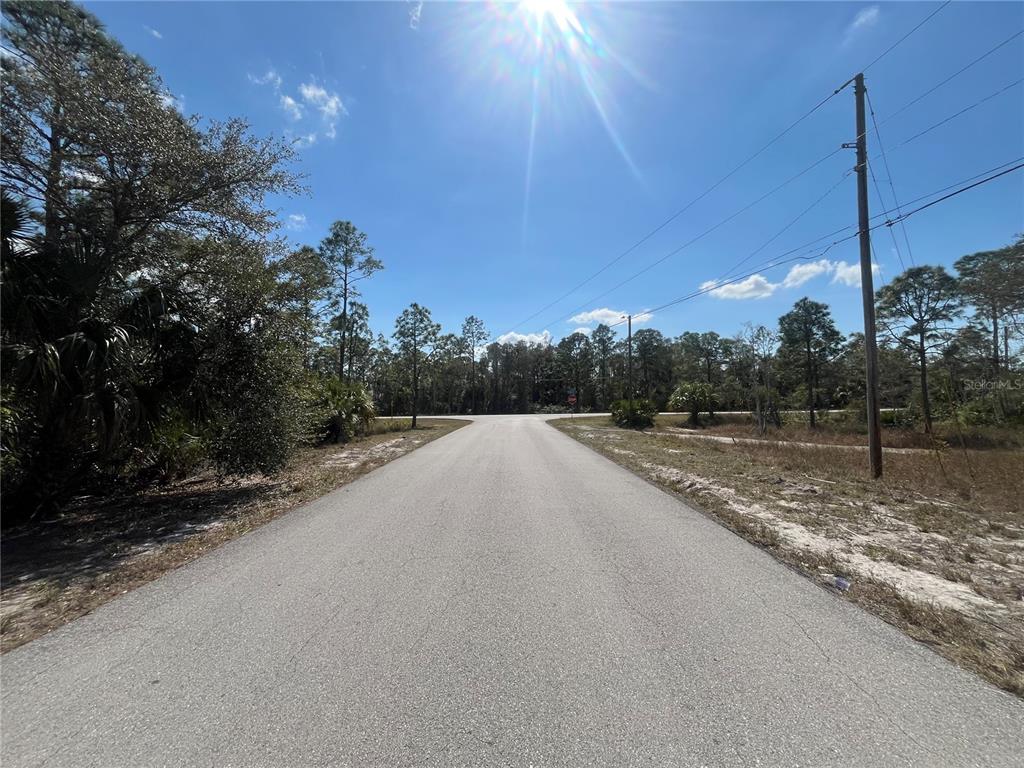 A paved road leads toward a T-intersection surrounded by trees under a bright blue sky — 494 BURRSTONE DRIVE, LEHIGH ACRES, FL