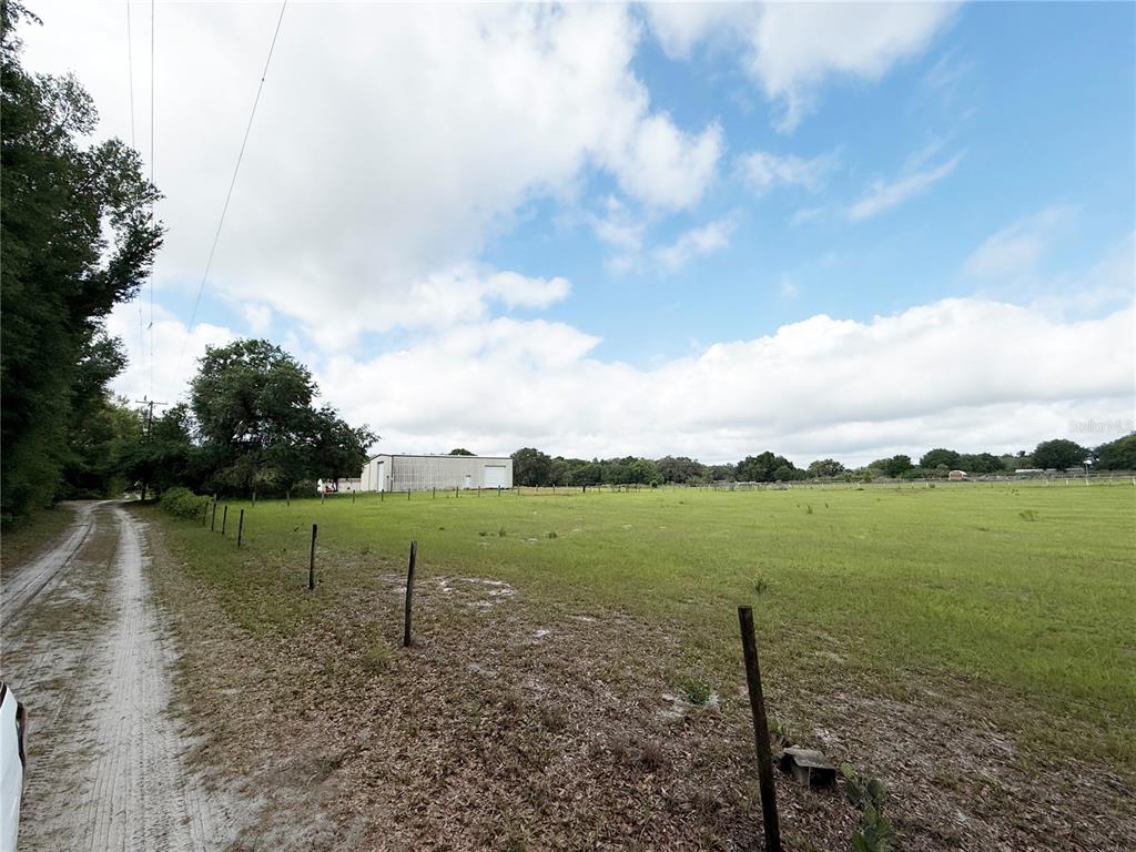 A dirt road runs alongside a large, fenced grassy field with a metal building beyond — SE SUNSET HARBOR ROAD, WEIRSDALE, FL