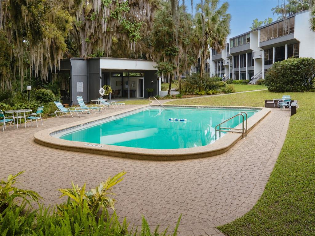 A rectangular swimming pool sits on a brick patio surrounded by trees and apartment buildings — 2818 SW 14TH DRIVE UNIT 107, GAINESVILLE, FL