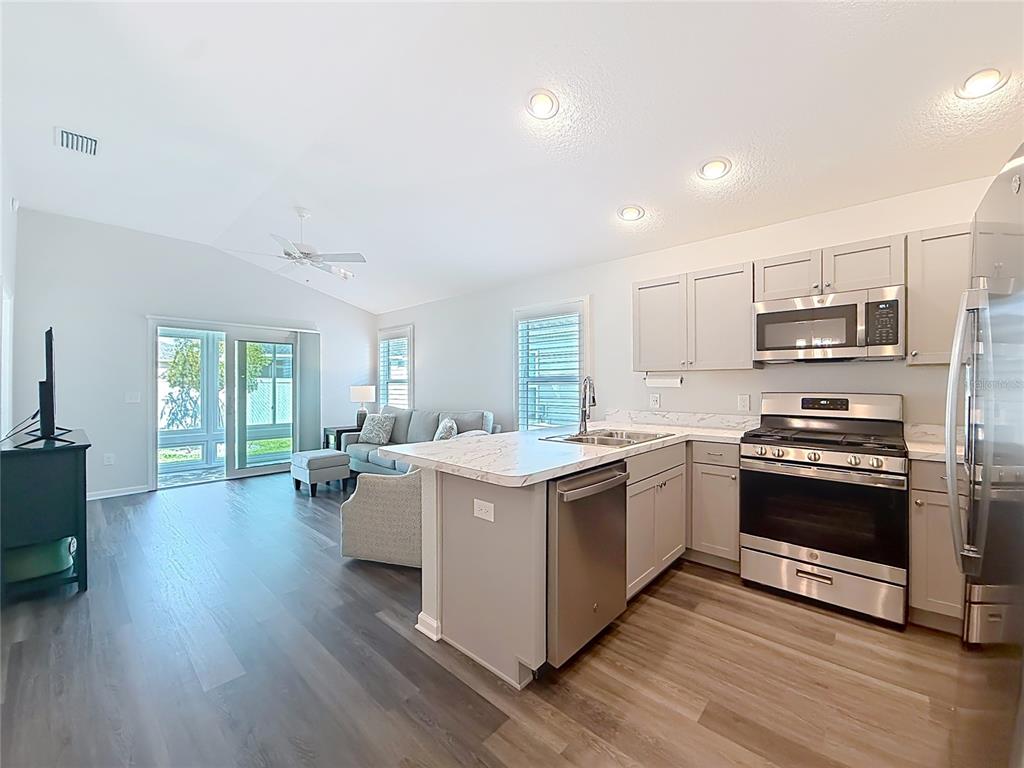 An open-concept kitchen with gray cabinets and stainless steel appliances overlooks a living area — 6966 LISA LANE, THE VILLAGES, FL