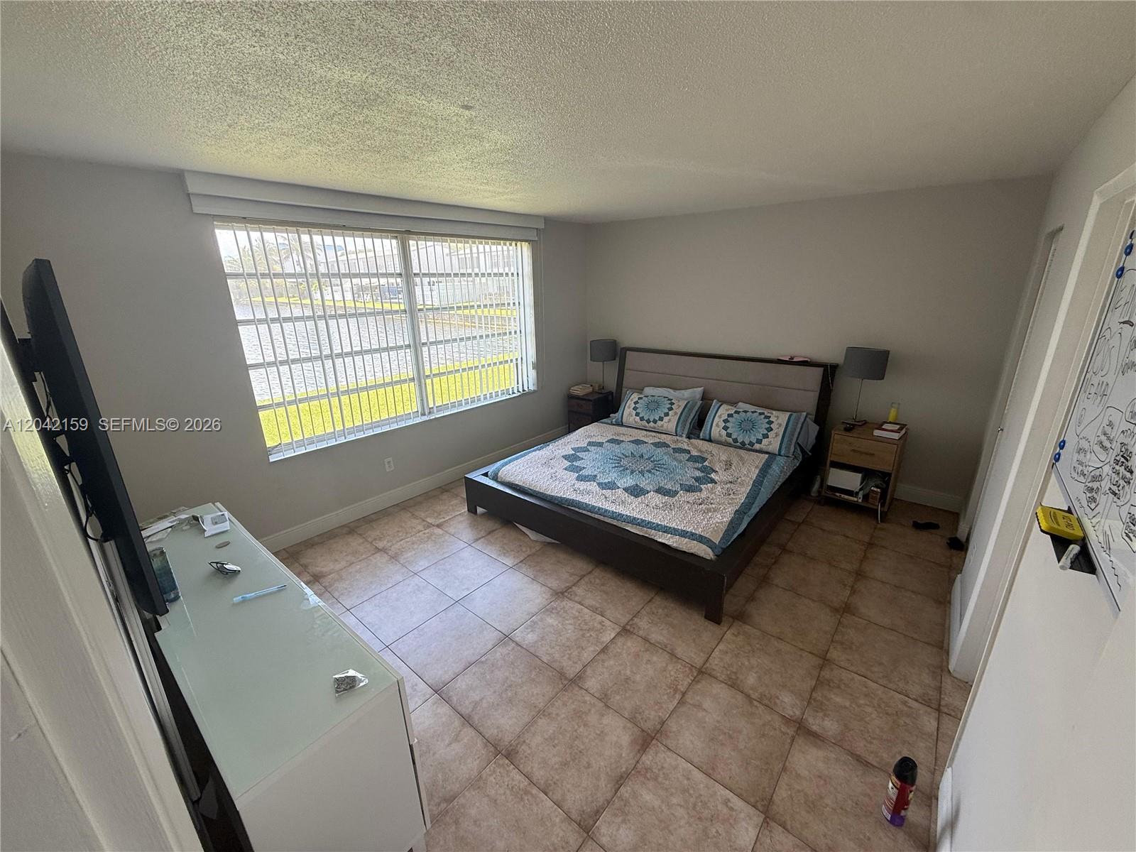 A bedroom features a bed with a patterned quilt, tiled flooring, and a large window — 1300 NE 191st St # A106, Miami FL 33179, MIAMI, FL