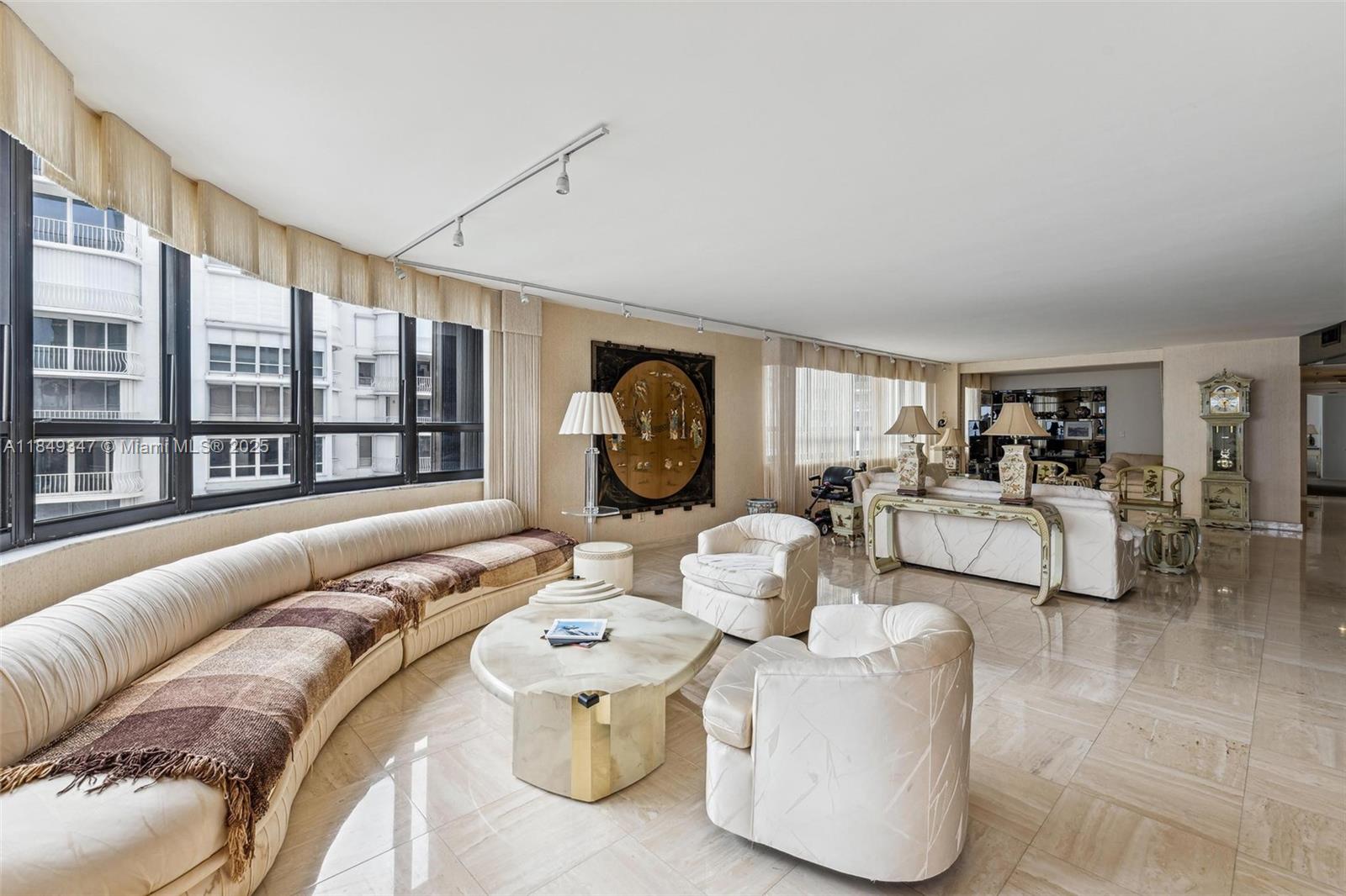 A spacious living room features a curved sofa, marble floors, and large windows with drapes — 10175 Collins Ave # 1202, Bal Harbour FL 33154, BAL HARBOUR, FL