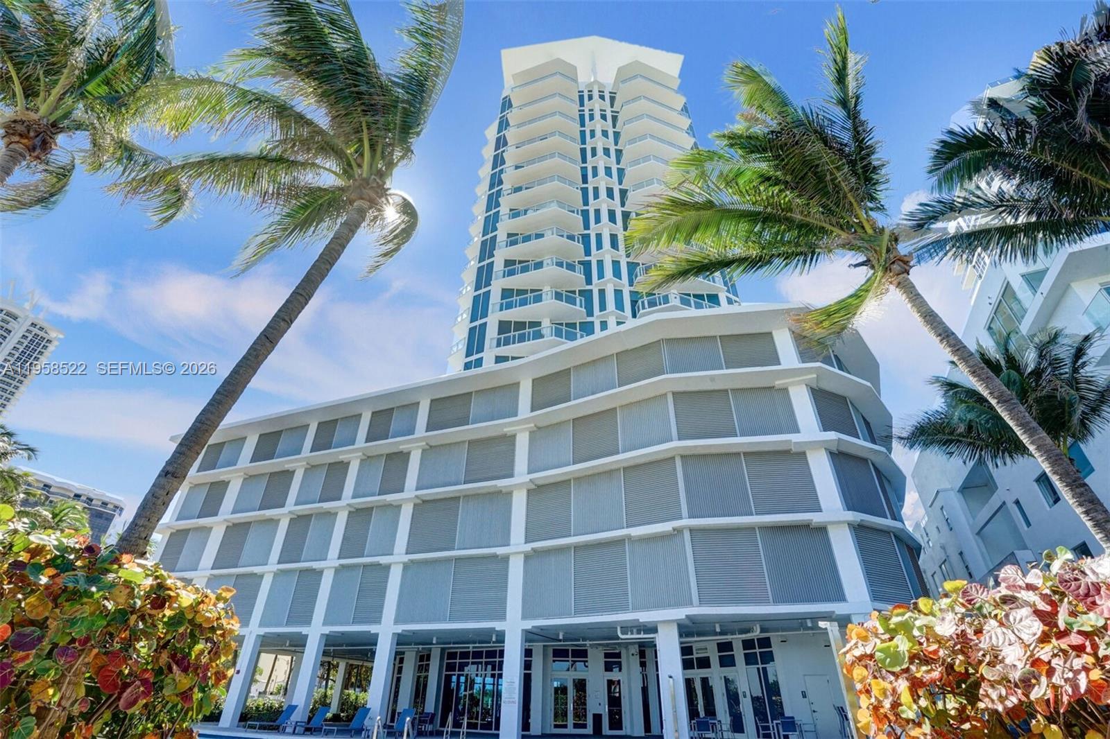 A tall white residential tower rises behind a multi-story building with louvered balcony shutters — 6515 Collins Ave # 901, Miami Beach FL 33141, MIAMI BEACH, FL